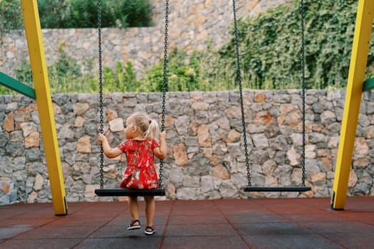 Little girl sits on a chain swing on a playground in the yard and looks away. Back view. High quality photo