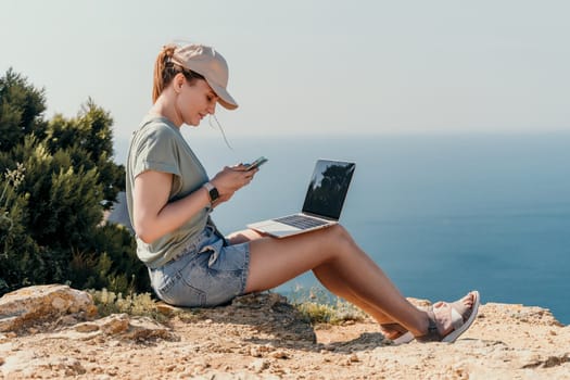 Digital nomad, woman in the hat, a business woman with a laptop sits on the rocks by the sea during sunset, makes a business transaction online from a distance. Freelance, remote work on vacation.