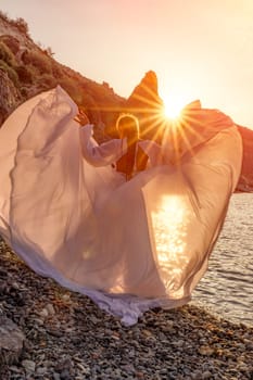 Mysterious woman silhouette long hair walks on the beach ocean water, sea nymph wind listens to the wave. Throws up a long white dress, a divine sunset. Artistic photo from the back without a face.