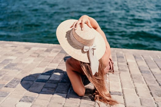 A woman in a swimsuit sits with her back holding a hat, looks at the ocean, sunny day, relaxes