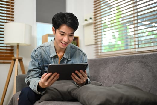 Smiling asian male freelancer sitting on couch and working with digital tablet.