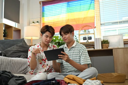 Young gay couple booking hotel reservation on digital tablet and packing luggage to travel vacation.