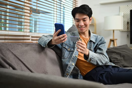 Satisfied young asian male holding credit card and shopping online on smartphone.