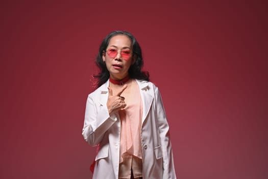 Cool and stylish middle age woman wearing pink glasses and fashionable clothes on red background.