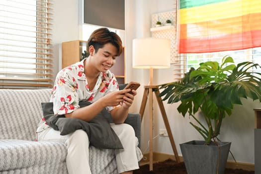 Carefree young gay male relaxing on couch and using mobile phone.
