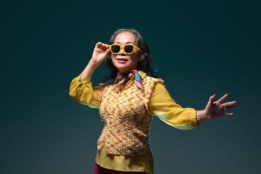 Stylish senior woman with fashionable clothes and sunglasses standing over green background.