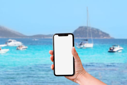 Hand holds mobile phone screen against of turquoise sea with yachts background.