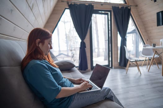 Red-haired woman typing on laptop while sitting on sofa in bungalow