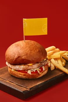 Tasty cheeseburger with chicken patty, tomato and lettuce in fluffy bun served with side of golden fries on wooden board, against bold red backdrop. Popular fast food dish