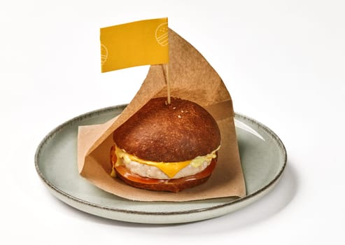 Tasty cheeseburger in fluffy browned bun with juicy chicken patty, slices of fresh tomato and cheese sauce in paper wrap with yellow label, presented on ceramic plate on white background