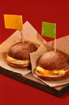 Two delicious burgers with juicy chicken and beef patties, tomato and cheese in browned buns decorated with colorful flags on toothpicks, served in craft paper on wooden serving board on red backdrop