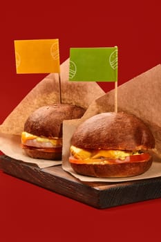 Two appetizing cheeseburgers in browned fluffy buns sprinkled with sesame marked with colorful flags with juicy chicken and beef patties, slices of fresh tomato in craft paper on wooden serving board