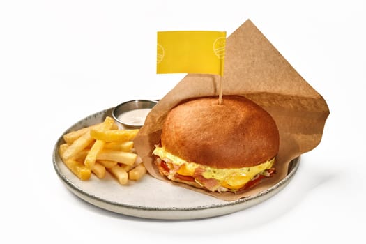 Delicious burger in white wheat bun with chicken patty, fried bacon slices, tomatoes and cheese sauce, wrapped in paper, served on ceramic plate with golden fries and spicy mayonnaise, on white