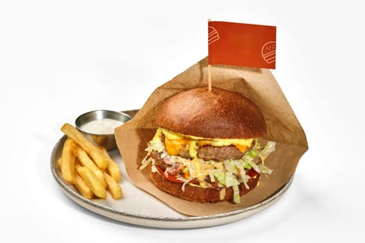 Appetizing hamburger in browned bun with two juicy patties, fresh tomatoes, lettuce and cheese sauce in craft paper wrapping served with french fries and spicy dip on plate, against white background