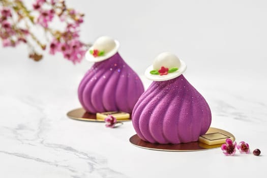 Chic velvety purple fig-shaped mousse confections topped by white chocolate hats, styled with delicate spring florals on marble table. Collections of signature handmade desserts