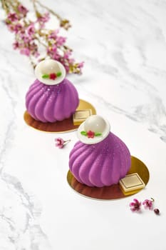 Sophisticated mousse pastries in vibrant purple with velvet texture, crowned with decorative white chocolate caps, set against backdrop of pink blossoms and marble