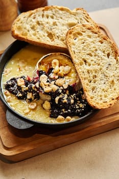 Baked creamy Camembert cheese topped with rich currant jam and crunchy hazelnuts, served with slices of toasted white spelt bread in cast iron skillet on wooden board