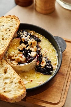 Creamy torched Camembert cheese in skillet, served with homemade sweet currant jam and hazelnuts, paired with crisp toasted slices of white spelt bread on wooden board