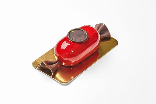 Luxurious artisan bonbon-shaped mousse dessert with glossy red glaze and embossed chocolate seal on gold tray, isolated on white background