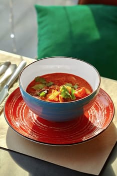 Bright and sunny dining setting with colorful ceramic bowl of fragrant savory Thai tom yam soup with seafood, shiitake mushrooms and cherry tomatoes garnished with fresh herbs. Traditional Asian dish