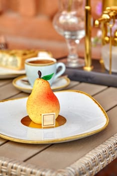 Tempting speckled yellow and red pear-shaped dessert with chocolate leaf and stem served on plate with cup of espresso on table in cozy cafe setting