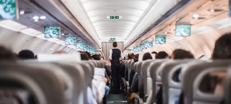 Interior of airplane with passengers on seats and stewardess in uniform walking the aisle, serving people. Commercial economy flight service concept