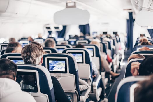 Interior of airplane with passengers on seats and stewardess in uniform walking the aisle, serving people. Commercial economy flight service concept