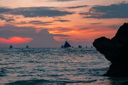 As the sun sets, the tranquil beach is bathed in soft hues of pink and purple. Boats with sails are visible on the horizon, gently drifting on the water. Boracay, Philippines.