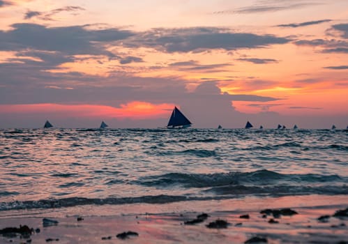 As the sun sets, the tranquil beach is bathed in soft hues of pink and purple. Boats with sails are visible on the horizon, gently drifting on the water. Boracay, Philippines.