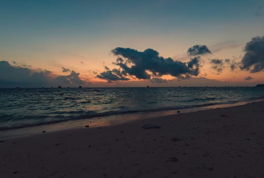 As the sun sets, the tranquil beach is bathed in soft hues of pink and purple. Boats with sails are visible on the horizon, gently drifting on the water. Boracay, Philippines.