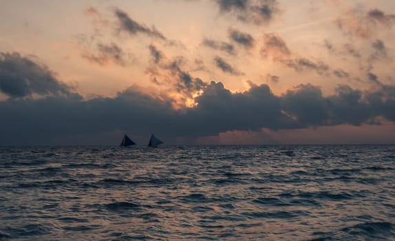 As the sun sets, the tranquil beach is bathed in soft hues of pink and purple. Boats with sails are visible on the horizon, gently drifting on the water. Boracay, Philippines.