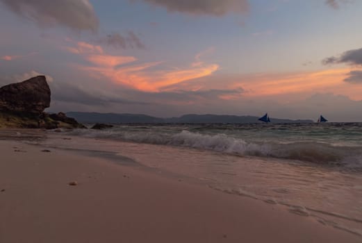 As the sun sets, the tranquil beach is bathed in soft hues of pink and purple. Boats with sails are visible on the horizon, gently drifting on the water. Boracay, Philippines.