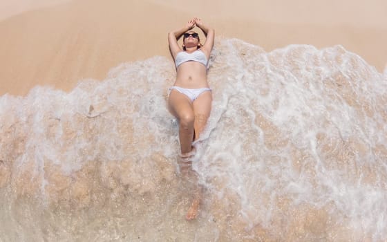 A woman lies on a sandy beach, her arms resting behind her head, as gentle waves lap around her. She is wearing a white bikini and sunglasses, enjoying the sunny afternoon at the shoreline.