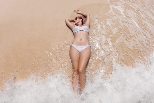 A woman lies on a sandy beach, her arms resting behind her head, as gentle waves lap around her. She is wearing a white bikini and sunglasses, enjoying the sunny afternoon at the shoreline.