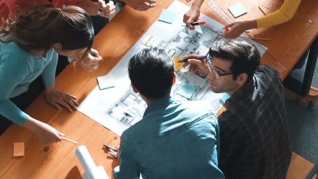 Top view of smart civil engineer working and writing blueprint while sitting at meeting table with blueprint, architectural equipment and colorful sticky note. Diverse team joining meeting. Symposium.