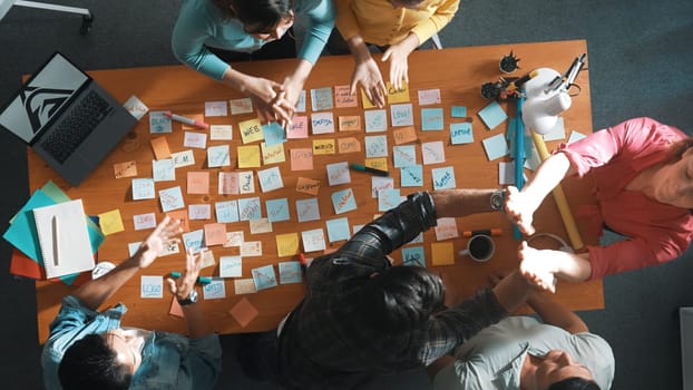 Top view of business team giving high five to celebrate project while standing at table with sticky notes and laptop. Aerial view of designer clapping hands to congratulate successful idea. Symposium.