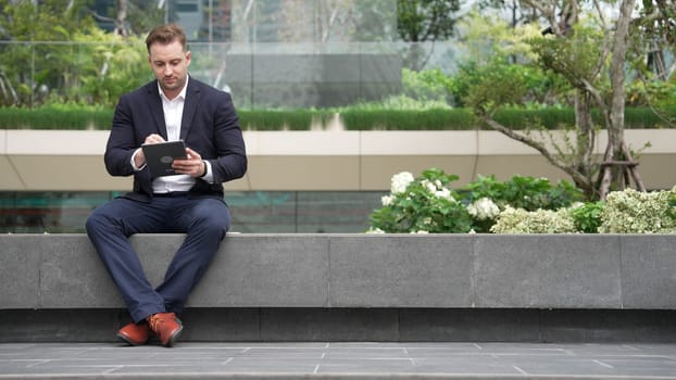 Caucasian smart businessman sitting at park while writing on tablet by using tablet pencil and planning marketing strategy. Manager looking at data analysis while thinking about financial plan. Urbane