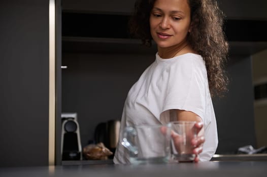 Mixed race beautiful smiling woman taking out a glass cup from a cupboard, putting it on the kitchen counter, standing in modern minimalist home kitchen interior. Morning routine. Healthy lifestyle