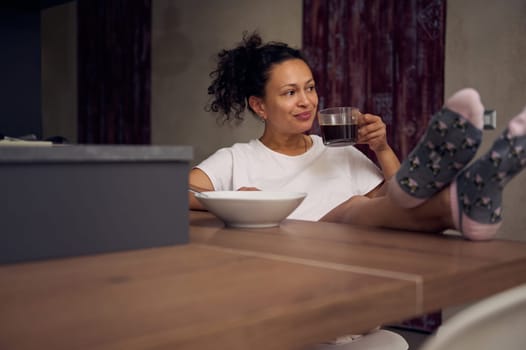 Pretty woman in pajamas, relaxing over cup of coffee. Happy young adult sitting at kitchen table, drinking hot coffee in the morning during her breakfast