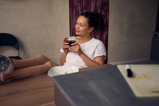 Pretty woman in pajamas, relaxing over cup of coffee. Happy young adult sitting at kitchen table, drinking hot coffee in the morning during her breakfast