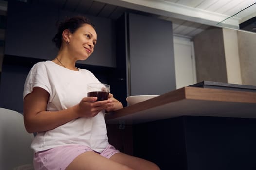 Pretty woman in pajamas, relaxing over cup of coffee. Happy young adult sitting at kitchen table, drinking hot coffee in the morning during her breakfast