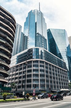 A towering modern skyscraper reflects the sky on its glass facade, surrounded by other tall buildings. Vehicles and a few people move along the bustling city street beneath.