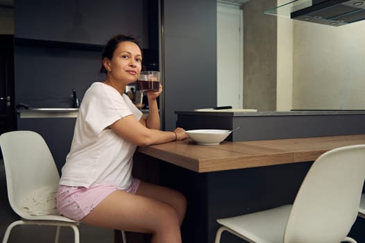 Pretty woman in pajamas, relaxing over cup of coffee. Happy young adult sitting at kitchen table, drinking hot coffee in the morning during her breakfast