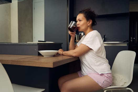 Pretty woman in pajamas, relaxing over cup of coffee. Happy young adult sitting at kitchen table, drinking hot coffee in the morning during her breakfast. People. Lifestyle