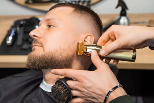 Barber cuts beard of young man making stylish shape with trimmer in barbershop closeup. Hairdresser serves male client in grooming salon