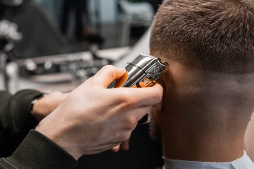 Barber uses shaver machine to cut young man hair in barbershop closeup. Master does modern haircut to male client in professional salon