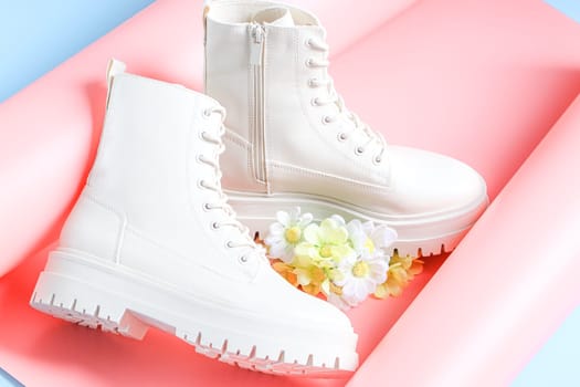 White demi-season boots made of eco-leather on a rough sole with lacing and a bouquet of spring flowers on a pink-blue background, flat lay close-up. The concept of fashion and women's shoes.