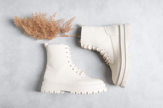 White demi-season boots made of eco-leather with clasps, laces, rough soles and a dry cane branch on a gray cement background, close-up flat lay. The concept of fashion and women's shoes.