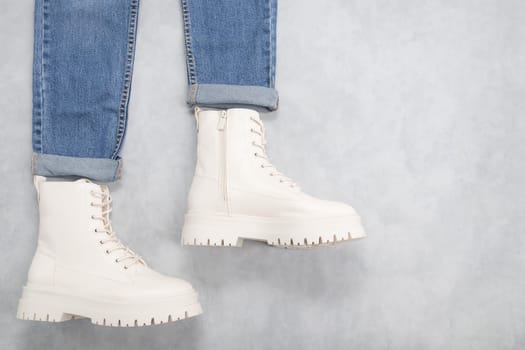 White demi-season boots made of eco-leather with fasteners, laces and blue jeans with a collar on the left on a gray cement background with copy space on the right, flat lay close-up. The concept of fashion and women's shoes.