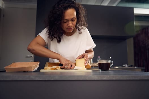 Beautiful multi ethnic young woman in white t-shirt, putting a slice of cheese over piece of bread, preparing sandwiches for snack